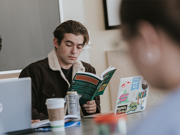 A student reads from a green book in a classroom. Open laptops, coffee cups and cans cover the table in front of him and another student, out of focus, sits near the front of the photo.
